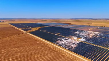 Açık mavi gökyüzünün altındaki güneş tarlaları. Geniş güneş çiftliği düz araziye yayılarak kırsal alanda enerji üretimi için güneş ışığını yakalıyor..