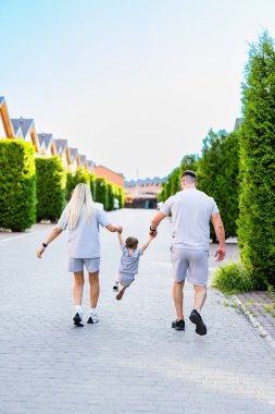 Aile gezintisi güneşte. Bir aile, neşeli çocuklarıyla el ele tutuşup güneşli bir günün tadını çıkarırken ağaçlarla kaplı bir sokakta yürüyor..