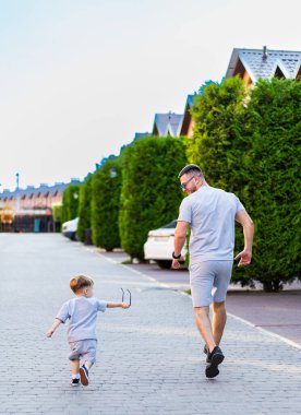 Baba ve oğlu buralarda geziniyor. Bir baba ve oğlu, arka planda yeşil çizgiler ve evler olan bir yerleşim sokağında geziniyor..