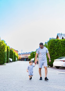 Baba ve oğul yaz güneşinde. Bir adam ve bir çocuk güneşli bir günde huzurlu, yeşil bir mahallede mutlu mesut yürürler..