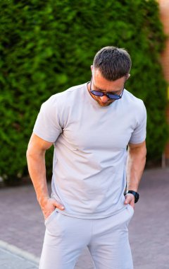 Gri takım elbiseli adam, sakin ol. Bir adam dışarıda duruyor, gri bir spor kıyafeti giyiyor, yeşil bir çalılıkta sakin bir ifadeyle aşağı bakıyor..