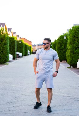 Genç adam buralarda geziniyor. Gri giyinmiş genç bir adam yeşil ağaçlarla çevrili sakin bir sokakta yürüyor..