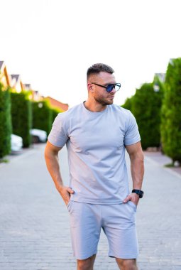 Güneşli bir günde günlük yaz tarzı. Açık gri elbiseli bir adam ağaçlarla ve evlerle dolu bir patikada güvenle duruyor..