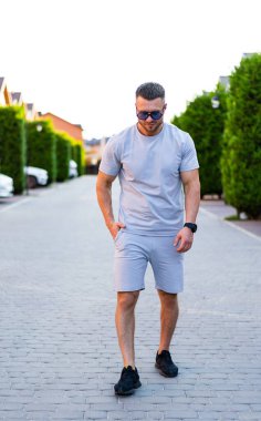 Yazın asfalt yolda yürüyen bir adam. Günlük kıyafetli bir adam ağaçlık bir caddede yürüyor, sıcak yaz akşamının tadını çıkarıyor..