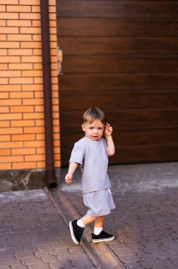 Kaldırımda tatlı bir çocuk yürüyor. Gri kıyafetli genç bir çocuk ahşap bir garaj kapısının yanındaki kaldırımda geziyor, güneşli bir günün tadını çıkarıyor..