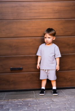 Garaj girişinde duran genç çocuk. Gri elbiseli genç bir çocuk ahşap bir garaj kapısının yanında duruyor, düşünceli ve yakınlardaki bir şeye odaklanmış görünüyor..