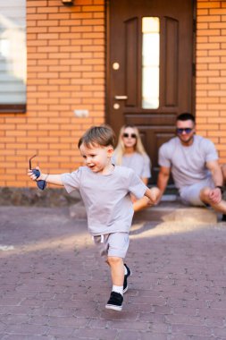Aile çocuğuyla dışarıda eğleniyor. Ailesi verandadan bakıp gülümserken, genç bir çocuk tuğla evin dışında neşeyle koşuyor..