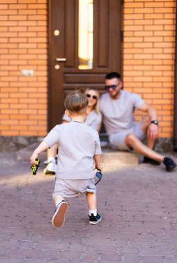Aile dışarıda eğleniyor. Günlük kıyafetli bir çocuk, güneşli bir günde evlerinin merdivenlerinde dinlenen ailesine doğru yürür..