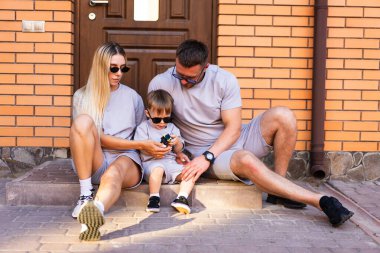 Aile dışarıda eğleniyor. Üç kişilik bir aile açık havada oturuyor günlük giysiler içinde, açık gökyüzü altında birlikte kaliteli zaman geçiriyorlar..