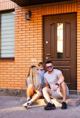 Dışarıda aile bağları. Üç kişilik bir aile evlerinin merdivenlerinde dinleniyor, gülümsüyor ve güneşli bir ortamda neşeli bir anı paylaşıyor..