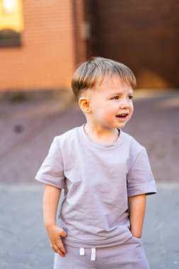 Çocuk dışarıda eğlenmekten zevk alıyor. Kısa saçlı genç bir çocuk gri bir kıyafetin içinde güneşli havanın tadını çıkarırken gülümsüyor..