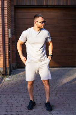 Rahat giyinen kendine güvenen bir adam. Bir adam rahat bir şekilde şort ve tişörtle duruyor, ahşap bir garaj kapısının yanındaki sıcak güneş ışığının tadını çıkarıyor..