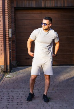 Sıradan kıyafetli bir adam evin dışında poz veriyor. Güneş gözlüğü takan bir adam evin dışında rahat bir şekilde durup güneşli bir günün tadını çıkarır..