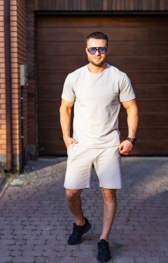 Hafif kıyafetler giymiş bir adam ahşap bir garajın yakınındaki kaldırım taşlı bir yolda geziniyor ve kendine güveni tam..