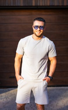 Adam güneşli bir günün tadını çıkarıyor. Gülümseyen bir adam, güneş gözlükleriyle rahat bir şekilde duruyor, ahşap bir garaj kapısının yanında güneşli bir günün tadını çıkarıyor..