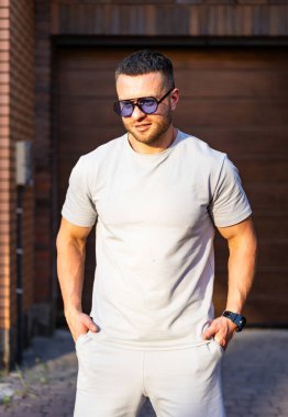 Dışarıda poz veren günlük kıyafetli bir adam. Bir adam özgüvenle garaj kapısına dayanmış, açık renkli tişört ve güneş gözlüğü takıyor..