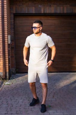 Sıradan yaz tarzında bir adam. Gri tişörtlü ve şortlu bir adam garajın dışında güneşli yaz havasının tadını çıkarıyor..