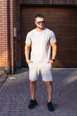 Gündelik giyimde kendine güvenen bir adam. Bir adam dışarıda taş bir yolda duruyor, açık renk bir kıyafet ve güneş gözlüğü takıyor, elleri ceplerinde..