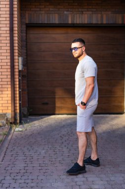 Adam şehirde dinleniyor. Genç adam güneşli bir günde, bir garajın yakınında şık bir kıyafetin içinde, kendine güven ve rahatlama yayıyor..