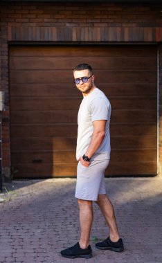 Sıradan kıyafetli bir adam garajın yakınında poz veriyor. Tişörtlü ve şortlu genç bir adam kahverengi bir garajın yanında duruyor, güneşli havanın tadını çıkarıyor..