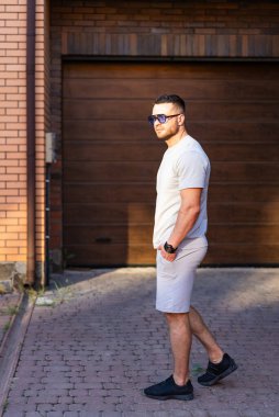 Güneşli bir günde günlük kıyafet. Bir adam, açık gri bir gömlek ve şort giymiş, gözlerini koruyan güneş gözlüğü takmış, güvenle kaldırımlı bir yolda duruyor..