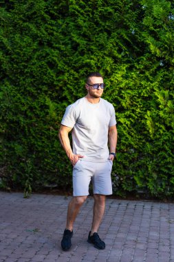 Yazlık kıyafetli kendine güvenen bir adam. Gri tişörtlü ve şortlu bir adam akşam vakti yemyeşil bir ortamda kendine güvenir..