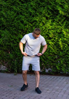 Adam yaz bahçesinde kıyafet ayarlıyor. Bir adam, yemyeşil yapraklarla dolu yemyeşil bir bahçede sıcak güneş ışığında dikilirken günlük kıyafetlerini ayarlıyor..