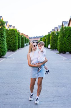 Güneşli bir yolda çocuğunu taşıyan bir kadın. Gündelik giyinmiş bir kadın çocuğunu açık bir gökyüzünün altında yeşil çitlerle kaplı güzel bir patika boyunca taşır..