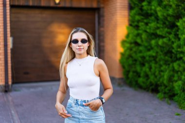 Genç bayan açık havada güneşli bir günün tadını çıkarıyor. Genç bir kadın, açık ve güneşli bir günde, güneş gözlüğü ve günlük kıyafetlerle, modern bir evin dışında güven içinde duruyor..