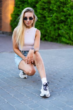 Dışarıdaki genç bayan günlük kıyafetlerle. Genç bir kadın kaldırımda diz çöker, güneş gözlüğü takar ve şık bir kıyafet giyer. Gün ışığında kendine güveni tamdır..