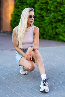 Kadın gündüz güven içinde poz veriyor. Genç bir kadın kaldırım taşlı bir sokakta diz çöküp yeşil yaprakların arasında şık kıyafetiyle ve güneş gözlüğüyle hava atıyor..