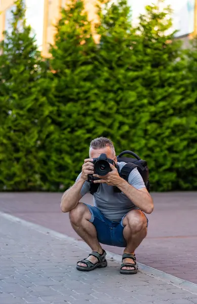 Fotoğrafçı park anlarını yakalar. Parkta çömelmiş bir adam kamerasıyla fotoğraf çekmeye odaklanmış. Yeşil ağaçlar doğal bir zemin sağlar.