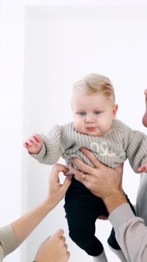 Gri kazaklı üç kişilik genç bir aile. Beyaz anne babalar küçük oğullarını yanaklarından tutup öperler. Beyaz zemin. Dikey video