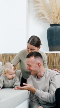 Mutlu ebeveynler kanepeye uzanmış şirin bebeklerinin yanına otururlar. Ciddi sarışın oğlan etrafa ilgiyle bakıyor. Dikey video