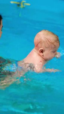 Esmer kadın, yüzme havuzunda bebek tutuyor. Sevimli çocuk suda yüzmek için ellerini hareket ettiriyor. Dikey video