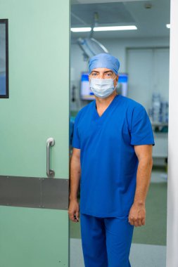 Cerrah ameliyathane girişinde. Ameliyat odasının kapısında duran mavi önlüklü ve maskeli doktor.