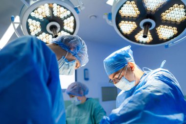 Cerrahlar ameliyat ışıkları altında ameliyat yapıyorlar. Mavi önlüklü cerrahlar modern bir ameliyathanede parlak cerrahi lambalar altında ameliyat yapıyorlar..