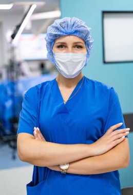 Kolları çapraz cerrahi önlüklü kadın doktor. Hastane koridorunda mavi önlük ve maske giyen kendine güvenen bir kadın doktor..