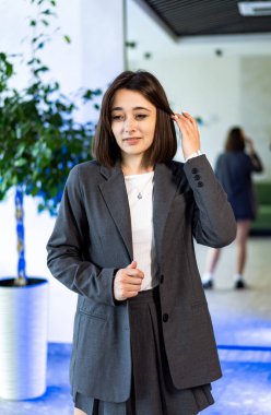 Resmi kıyafetli genç bir kadın içeride saç ayarlıyor. Gri takım elbiseli genç bir kadın saçlarını aynanın ve yeşil bitkinin yanında dikilirken düzeltiyor..