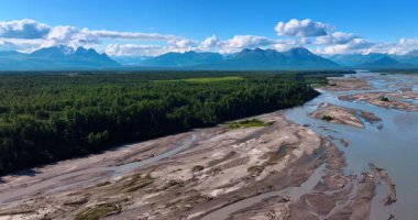 Yeşil yemyeşil ağaçlarla dolu sığ bir nehir. Güzel pofuduk bulutlar mavi gökyüzünde asılı arkaplanda dağ sırasının üzerinde.