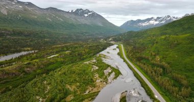 Cars go by the highway along the shallow river flowing in the valley. Spectacular mountains with some snow on top at backdrop. Alaska, USA.