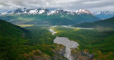 Nehir kıyısındaki dağlardaki güzel yeşil vadiye yaklaşıyoruz. Kar kaplı kayaların üzerindeki gri bulut. Hava görüntüsü. Alaska, ABD.