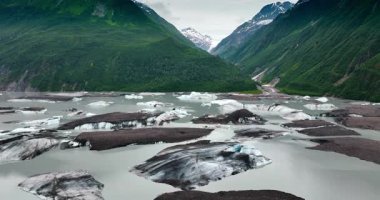 Ice lumps sticking out of grey riverscape. Huge verdant mountains on the waterfront at backdrop. Alaska, USA.