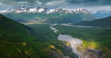 River flows among the green woods in the vast valley. Drone footage above the majestic scenery of Alaska approaching the snow-capped mountains. Aerial view.