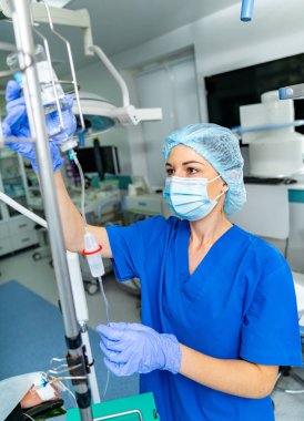 Kadın hemşire ameliyathanede serum sistemini ayarlıyor. Mavi önlüklü ve maskeli bir tıp hemşiresi ameliyathaneye damar yolu açıyor.