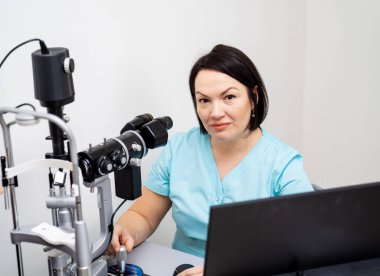 Klinikteki Slit Lambası 'nda oturan kadın göz doktoru. Göz teçhizatıyla çalışan turkuaz üniforma uzmanı..