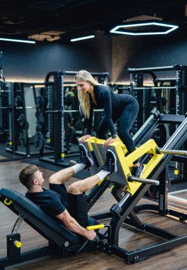 Sporcular fitness kulübünde bacak pres makinesi kullanıyorlar. Kadın modern spor salonunda onu desteklerken erkek bacak pres makinesinde egzersiz yapıyor..