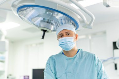 Ameliyathanede tıbbi maske takan bir cerrah. Cerrahi önlüklü ve maskeli bir erkek doktor hastanede ameliyat lambasının altında duruyor..