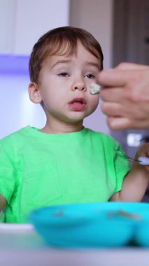 Lovely dark-haired baby boy having meals. Kid sits at the high chair in the kitchen with spoon in hand. Low angle view. Vertical video