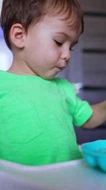 Mom's hand shoves the full spoon to her son. Baby boy refuses mom's spoon but takes some food with his own spoon. Low angle view. Vertical video